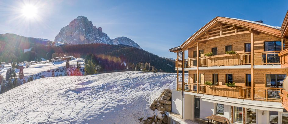 Exterior shot of the catered ski chalets of the Hotel Rodella on a sunny day with snow, forest and mountain in the background.