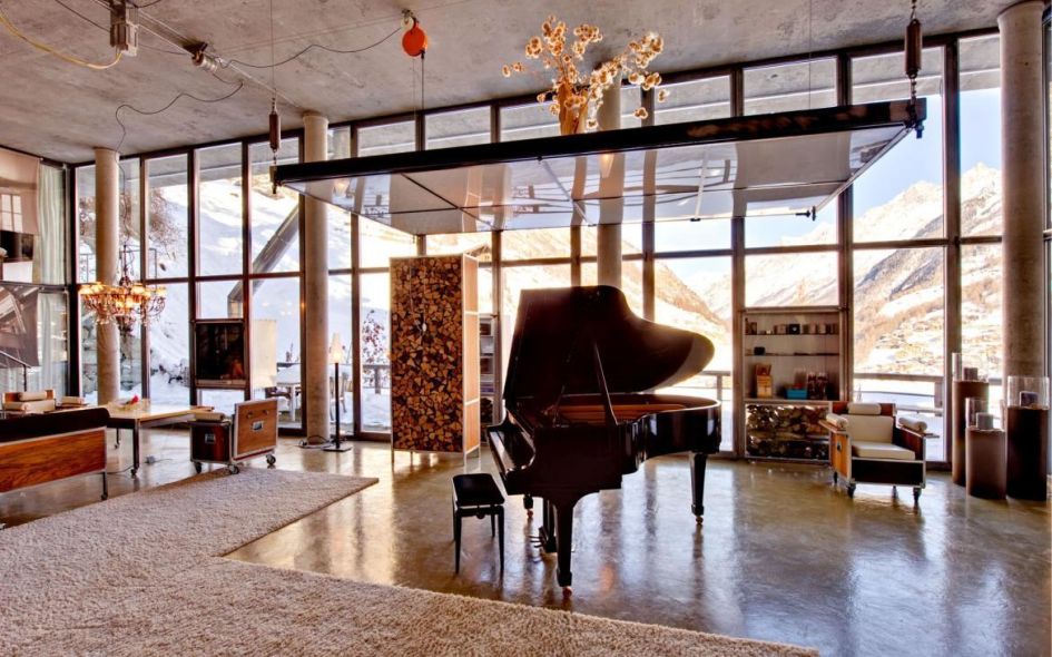 Pictured is the grand piano in the living room of Heinz Julen Loft, Zermatt, with a backdrop of the Mischabel mountains through the windows. A luxury chalet feel throughout.