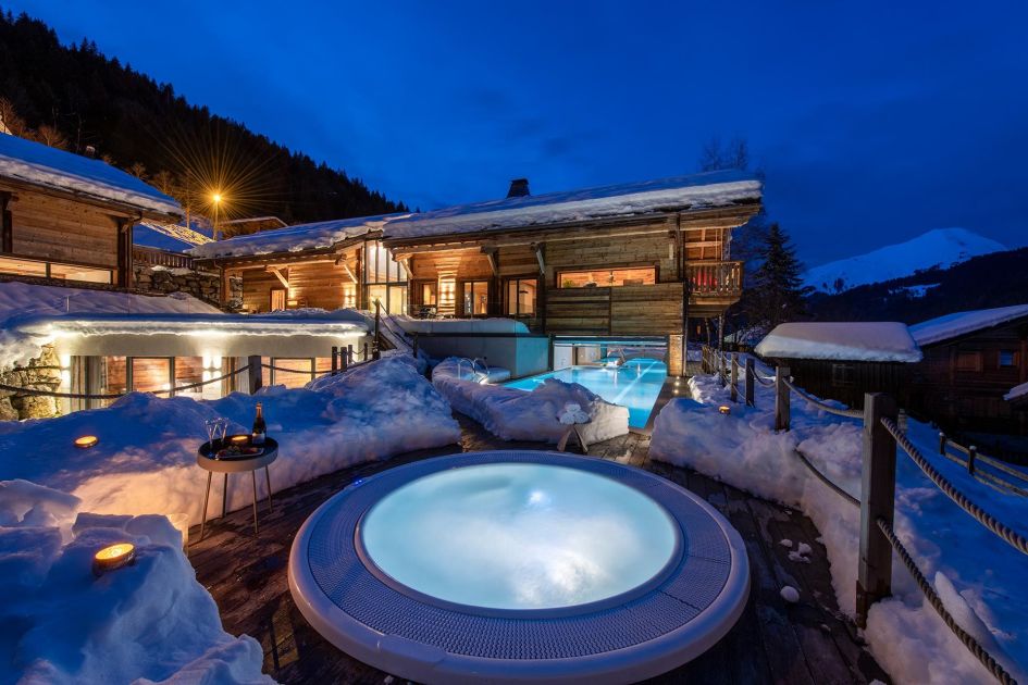 Outdoor sunken hot tub on the terrace of Chalet Joux Plane in Morzine. Nighttime shot with the cosy chalet and indoor/outdoor pool in the background. 