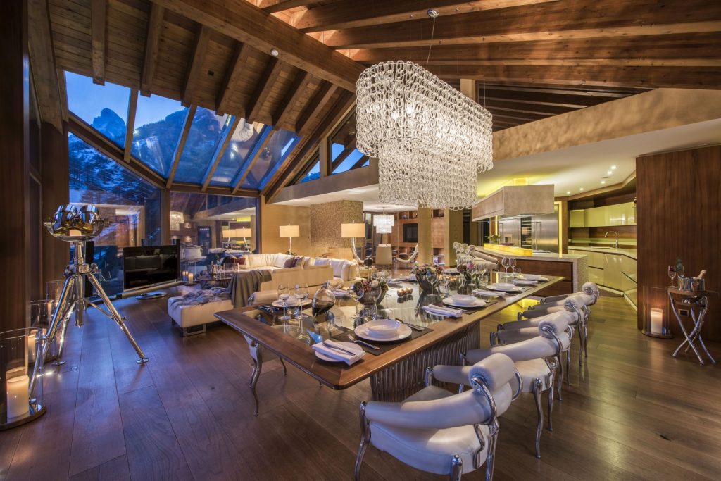 A picture showing the stunning chalet interior of Chalet Zermatt Peak, featuring the dining room and lounge. The chandelier is impressive, and the Matterhorn views are visible in the background.