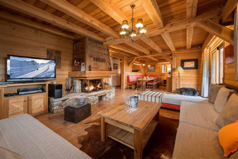 Chalet Le Manoir is a beautiful alpine chalet in Alpe d'Huez with an open-plan  living space, complete with traditional wooden cladding, wood-burning fireplace, sofa corner and dining table in the background.