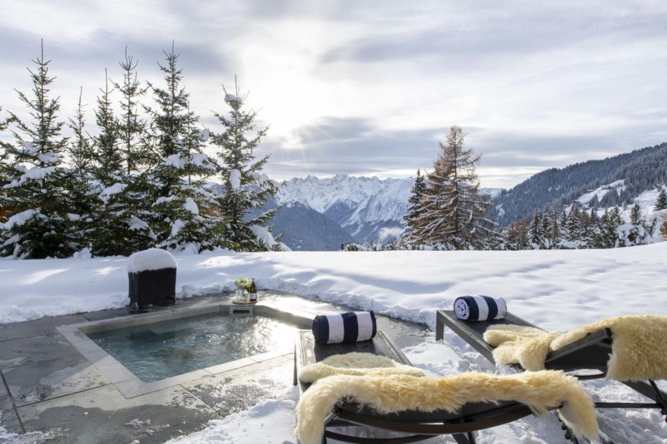Outdoor sunken hot tub at Chalet Teredo with the privacy of snow-covered trees and the advantage of snow-capped mountains. Loungers draped with fluffy blankets complete the scene for the perfect setting post skiing in Verbier 