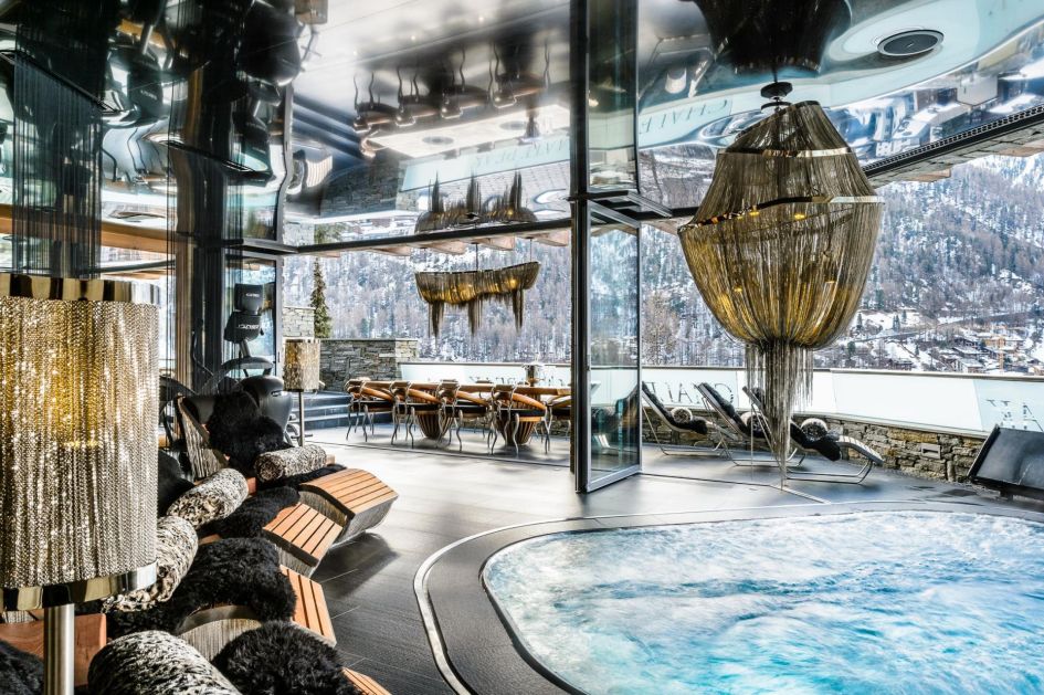Indoor hot tub at Chalet Zermatt Peak surrounded by loungers and embellished with grand lighting fixtures and top quality interior design. There are sliding doors for the effect of an indoor/outdoor feel. 