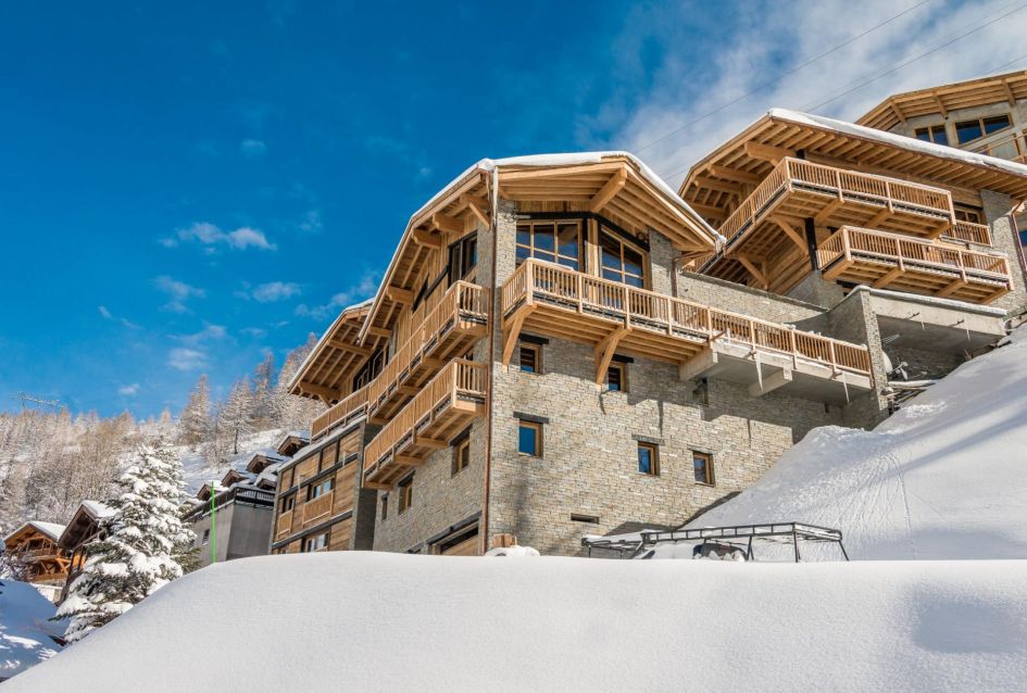 Exterior picture of The Black Diamond residence featuring a light stone and wooden facade, snow in the foreground and blue skies above - a great example of why you should consider a mountain residence for your luxury ski holiday!