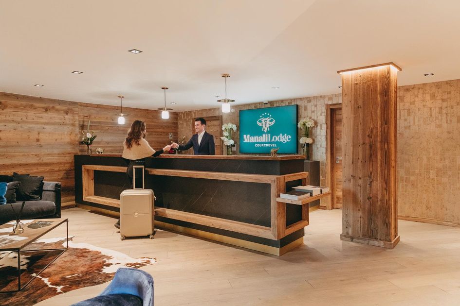 A man stood behind a black reception desk, handing a woman keys; the room is decorated in alpine wood. A blue sign reading 'Manali Lodge Courchevel' is at the back behind the desk.