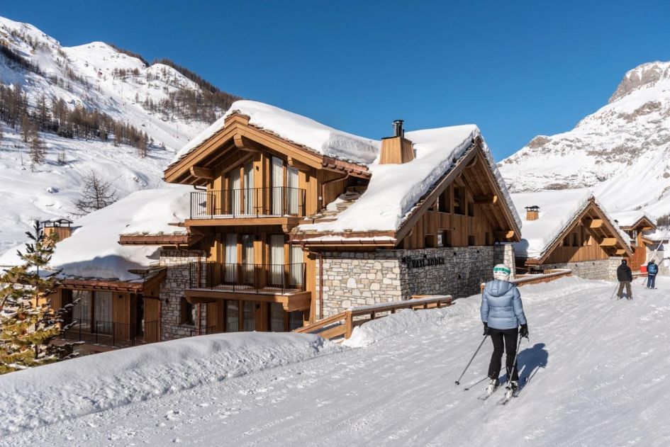 The exterior of Vail Lodge Residence in Val d'Isere with snowy mountain views in the background and skiers passing by the front of its wooden and stone building. Close to the centre of Val d'Isere, the exposed stone and wooden building lies in an exceptional ski in, ski out location!