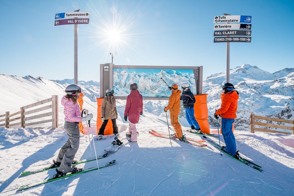 A group inspecting a piste map on the mountain, deciding where's best to ski in Tignes. Blue skies and snowy views feature behind the map. Ski residences offer great options for group ski holidays. 