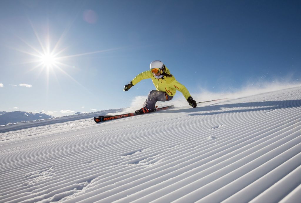 Skier carving on a groomed piste with blue skies and sun behind.