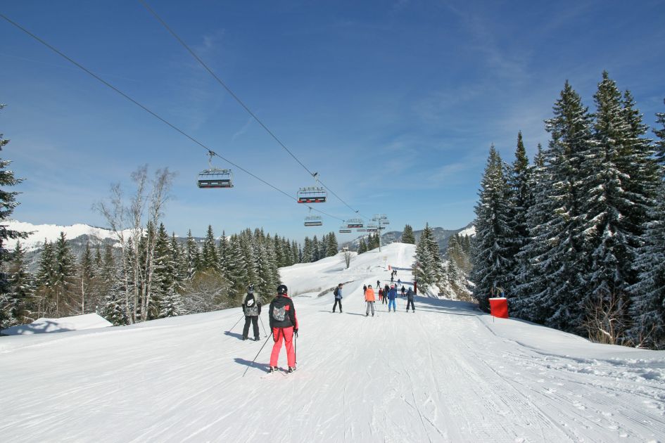Skiers on a piste in Morzine, under blue skies and a chair lift. Trees line the edge of the run, lightly dusted in snow.