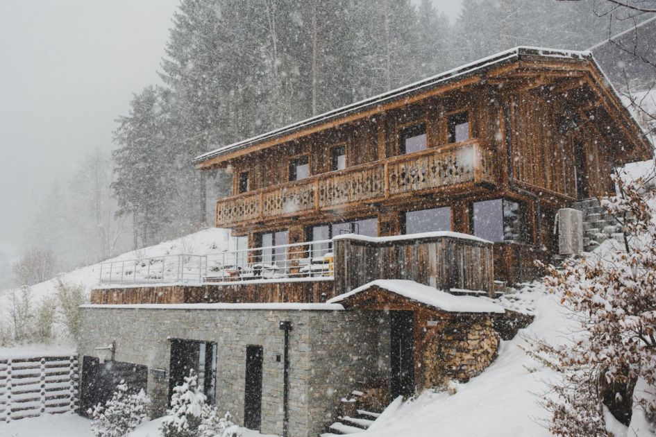 Alpine Retreat, a luxury chalet in Morzine, shows its wooden exterior pictured in a snow blizzard. Snow can be seen falling against the chalet's wood, whilst also being surrounded by snow.