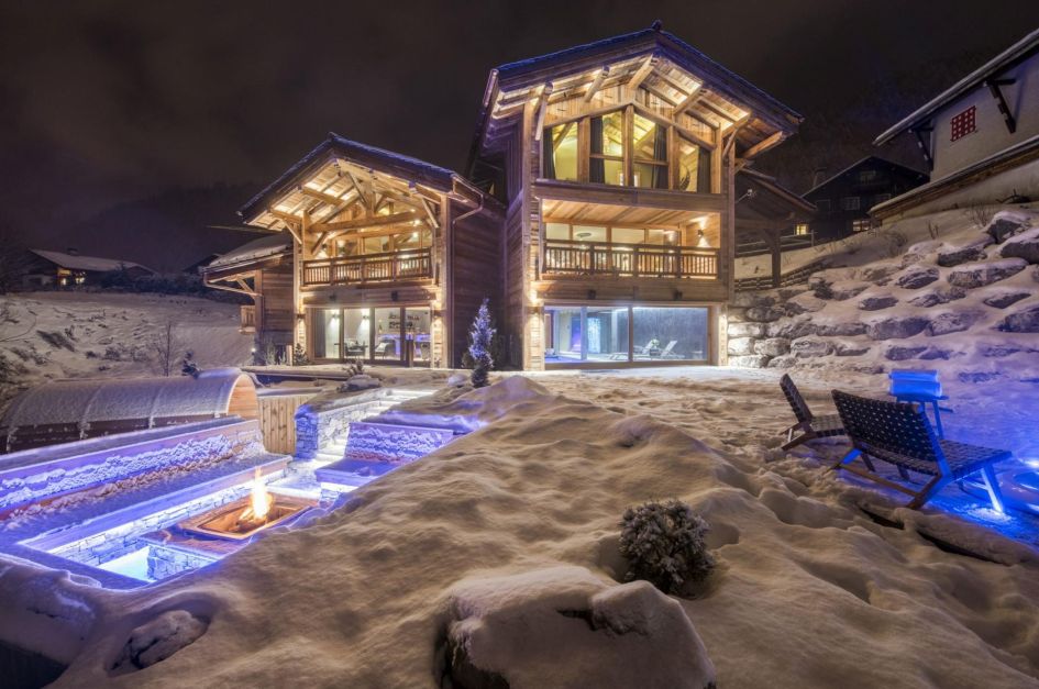 A well-lit picture of Chalet Sapphire's terrace lit up at night, surrounded by snow, with the chalet buildings, firepit, barrel sauna and outdoor seating all on show.