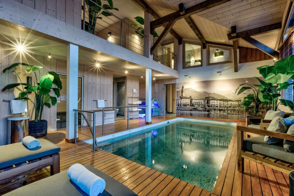 Picture of Chalet X's spa area, featuring an indoor swimming pool in the foreground. Decorated in light alpine woods and having high ceilings, the loungers on either side of the pool make for a great spot to relax.