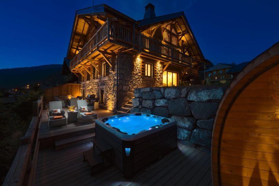 Exterior photo taken from The Old Macaroni's terrace at night. The corner of a barrel sauna is visible, and the outdoor hot tub, with the well-lit chalet showing the traditional stone exterior in the background.
