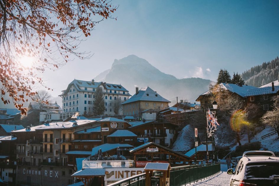 Staggered traditional and quaint buildings in Morzine, with a road to the right side of the image. In the background, the Nyon mountain pokes above the buildings.