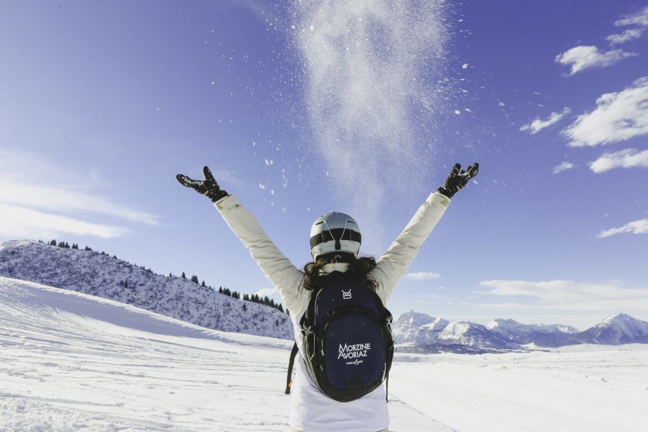 Woman throwing snow in the air whilst blue skies and mountains feature in the distance. A picture from Morzine ski resort.