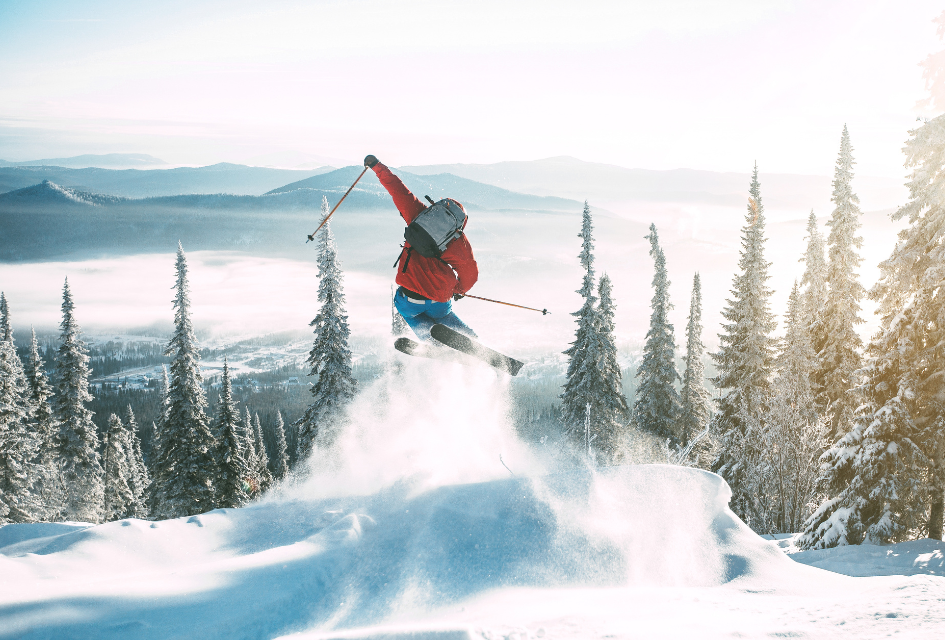 Person skiing. Credit: Kirill Lazarev via Canva photos A person from behind skiing off a jump surrounded by a plume of snow with mountains and trees in the background, enjoying the benefits of the Ikon and Epic Passes.