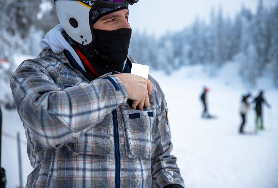 Ikon Vs Epic Pass. Credit: PhotoLife94 via Canva.com Man putting a ski pass into front pocket of jacket, with the snow and trees behind.