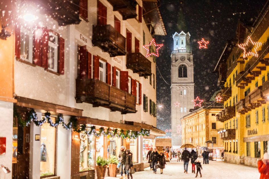 The famous Corso Italia, the main high street through Cortina, in snowy weather at nighttime with the iconic church in the background.