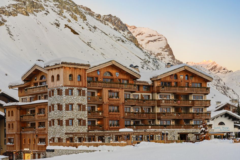 One of the best luxury ski hotels in Europe for location, the stone and wood exterior of Hotel Airelles Val d'Isère is seen here adjacent to the snow front in resort, with mountains in the background. 