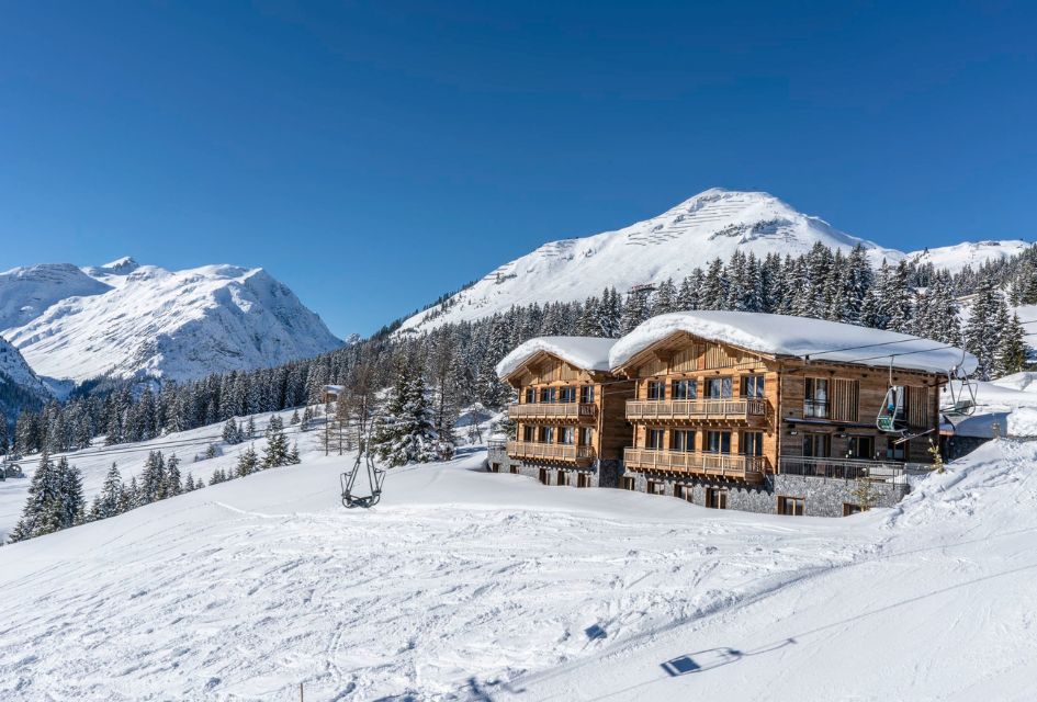 Two luxury ski-in, ski-out chalets in Lech with a chairlift passing it by and snowy moutnains in the background. 