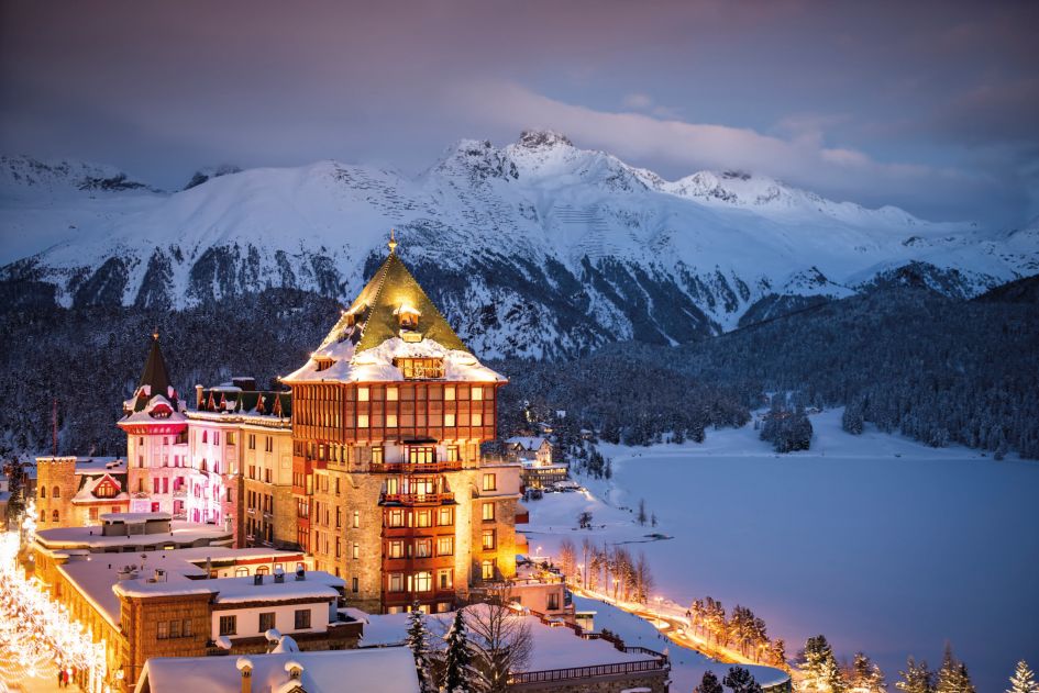 The lit up exterior of one of the oldest and best luxury ski hotels in Europe, Badrutt's Palace, at dusk. The frozen Lake St Moritz sits next to it, with mountainous views in the background. 