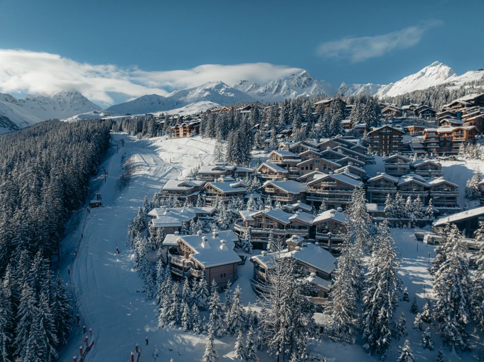 One of the best luxury ski hotels in Europe for ski in/ski out access, this image shows a drone shot of the various chalets and chalet-style buildings that make up the Hotel Le K2 Palace, sat next to the blue Cospillot piste on a sunny day in Courchevel 1850.