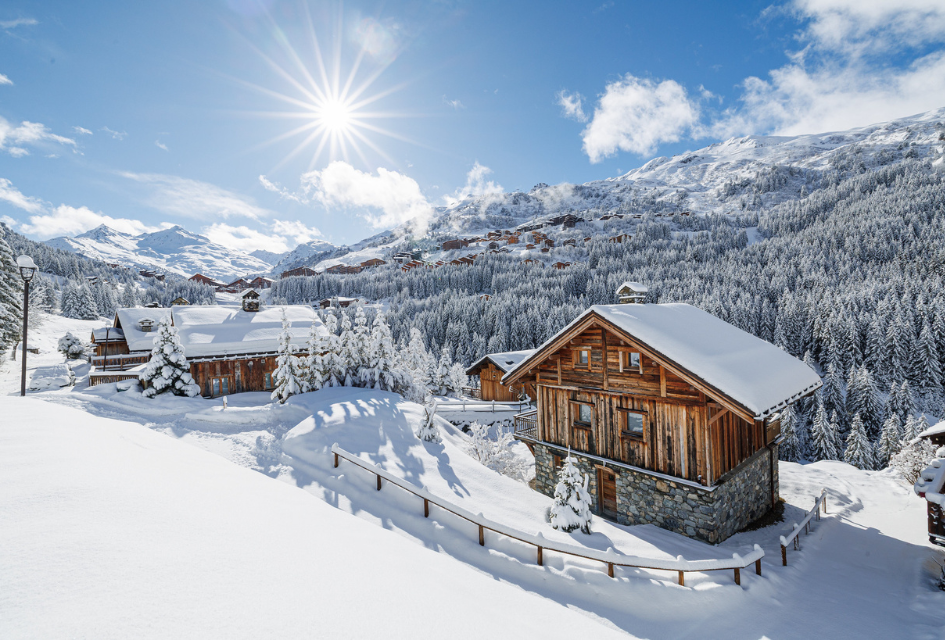 View of Meribel with snow covered chalets and tree-covered mountains in the background. The sun shining down on a crisp winter's day. 