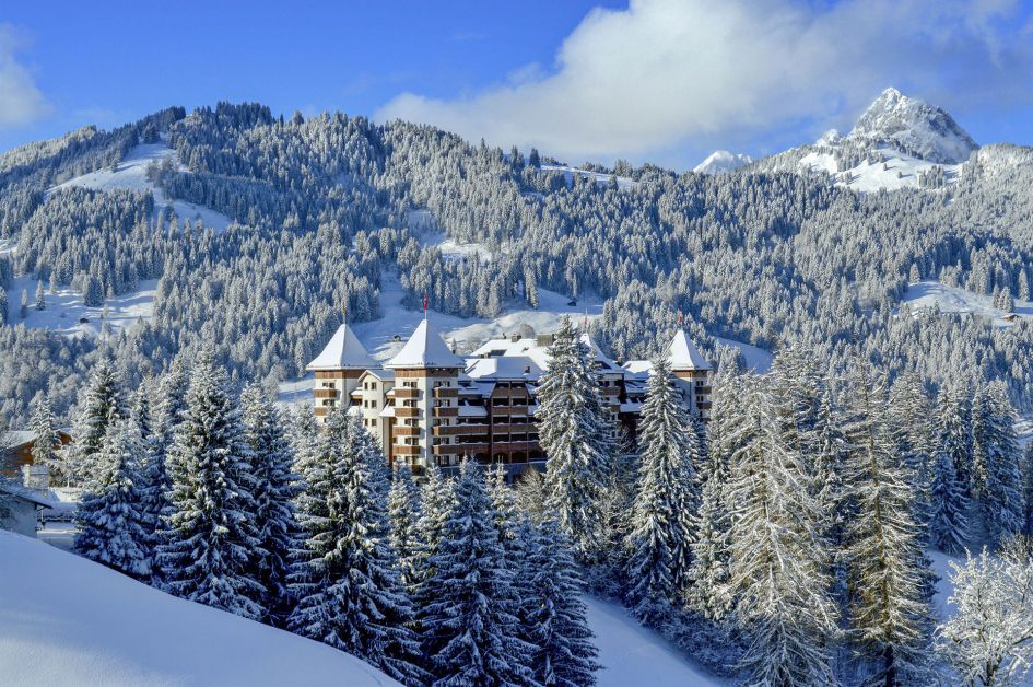 The corner towers, roof, and part of the exteriors of the Alpina Gstaad hotel, nestled amongst trees and mountains, all of which have been blanketed by snow, on a blue sky day.