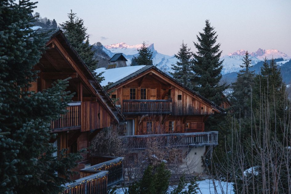 Close up of the wooden exterior of the Alpaga Hotel in Megève, with fir trees surrounding and snowy mountain peaks in the background. 