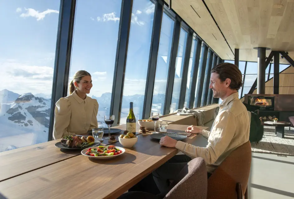 Dining at Cabane Tortin Couple enjoying dinner and wine at the dining table of Cabane Tortin, fire roaring in the background and mountains on view from the huge windows.