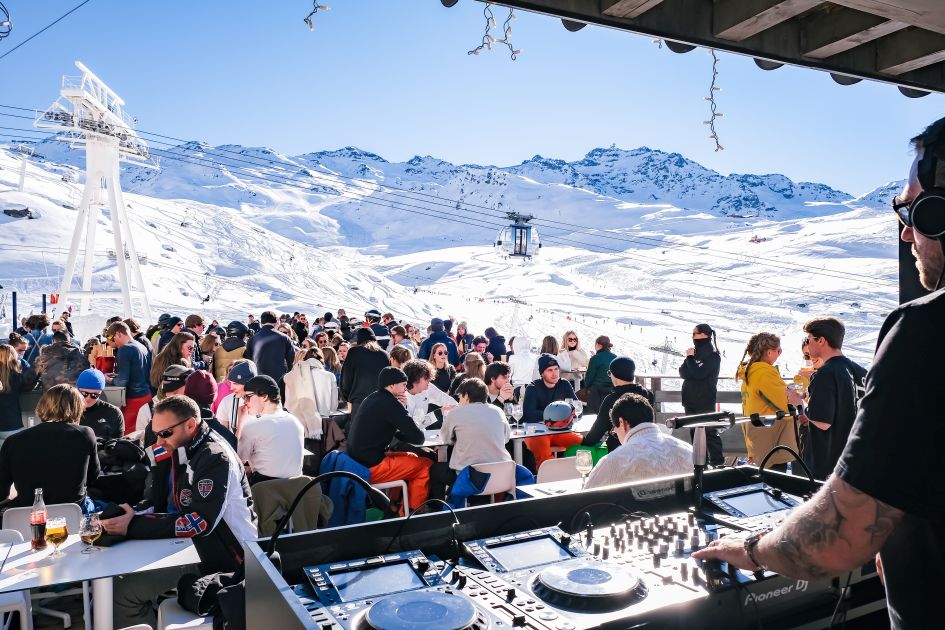 Après ski in Val Thorens on the terrace of Fahrenheit 7. A DJ is in the foreground, with people sat with drinks on the terrace and the mountains and gondola in the background.