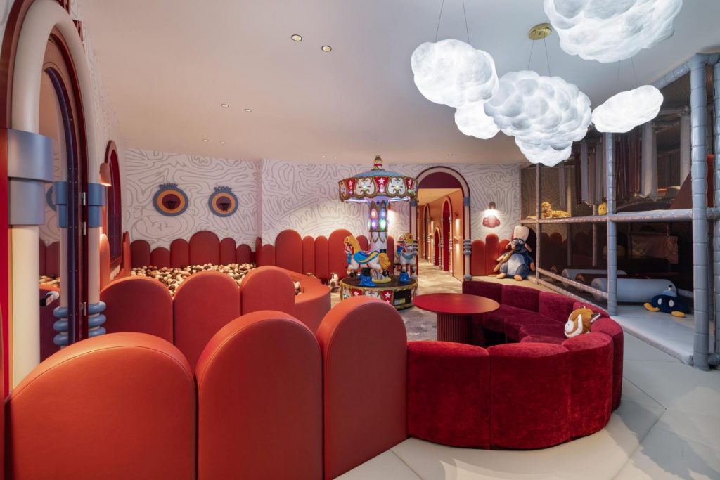 Le K2 Kids Club Kids club in Hotel Le K2 Palace, featuring velvet seating with whimsical cloud shaped pendant ligthing installations. The room hosts an indoor ball pit and a central decorative carousel ride.