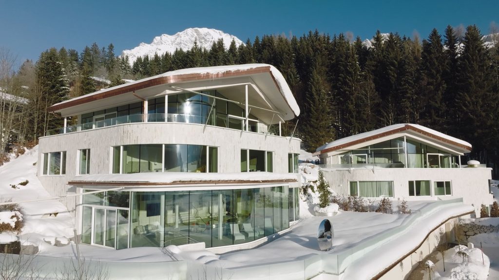 Julisam Estate, Leogang Julisam Estate built into a snow-covered hillside, set against a backdrop of a dense pine forest and a towering, snow-capped mountain peak under a clear blue sky.