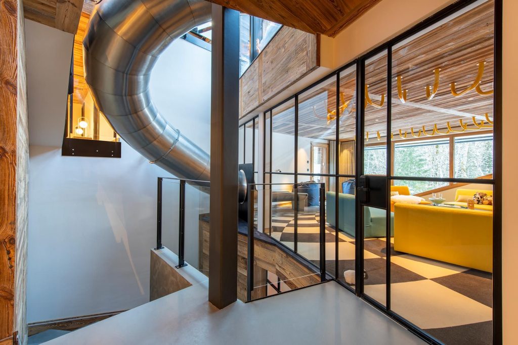 Chalet Tataali, Morzine A slide leads from the floor above into a vibrant lounge and playroom space featuring distinctive checkered flooring, bright yellow contemporary seating with monkey bars ahead